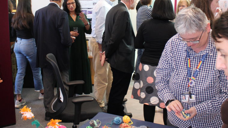 A group of people look at scientific research posters and chat. In the foreground are some brightly coloured pom-poms to demonstrate a scientific engagement activity,